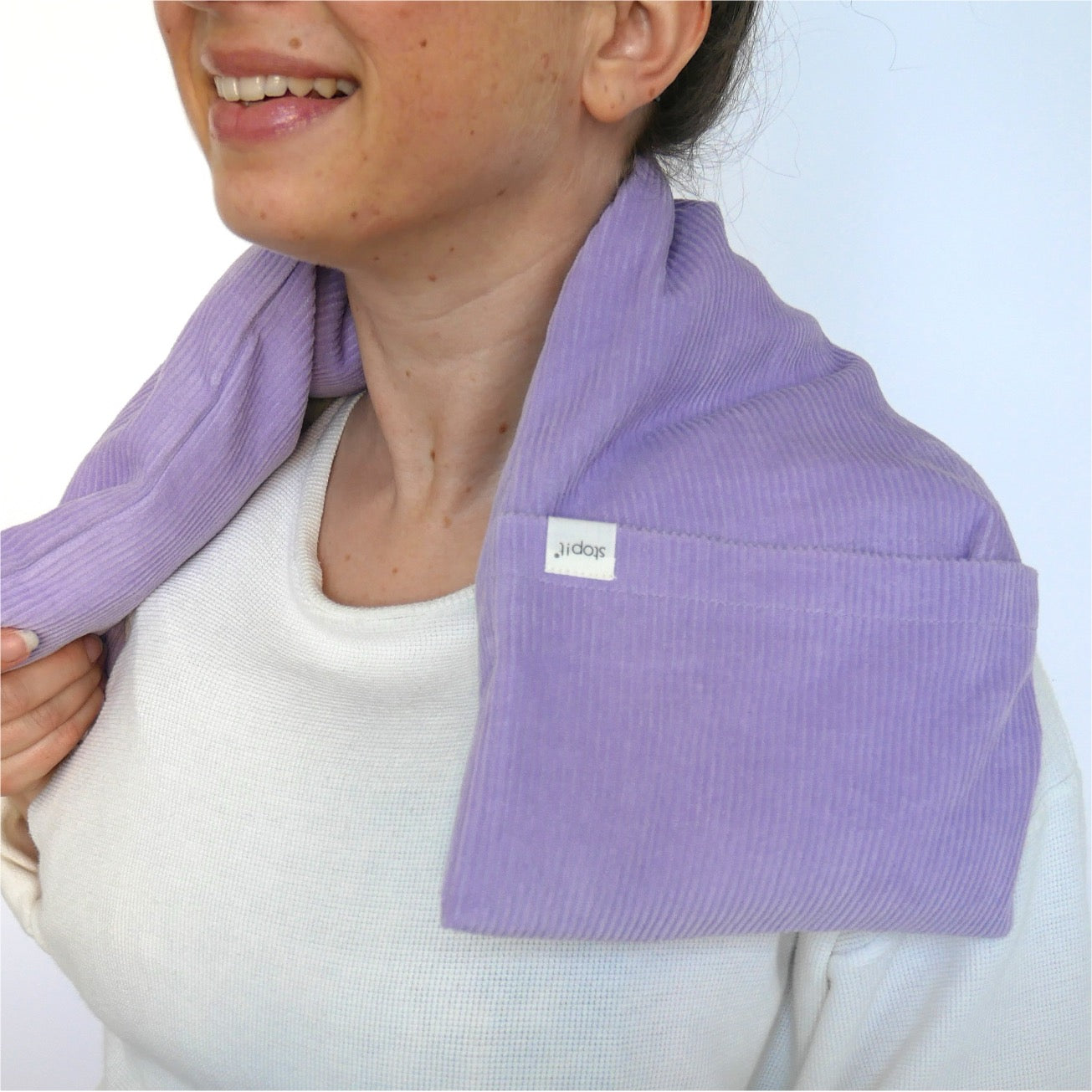 Wärmekissen Hot Cordie Lilac Tree liegt um den Nackenbereich einer lächelnden Frau, die schräg von vorne im Bild ist. Sie ist von der Nasenspitze bis unter die Brust zu sehen und trägt ein helles Shirt.