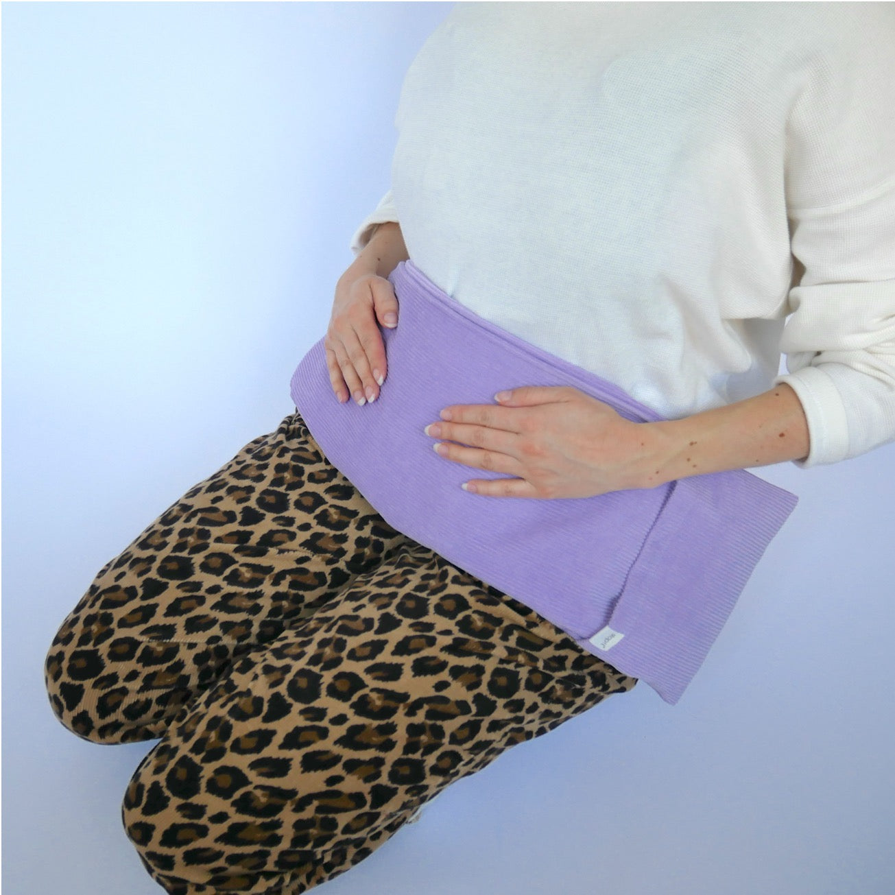 Wärmekissen Hot Cordie Lilac Tree liegt um den Bauchbereich einer Frau, die schräg von vorne im Bild ist. Sie ist von der Schulterpartie bis einschließlich zu den Beinen zu sehen und trägt ein schwarzes Shirt und eine Hose mit Leopardenmuster. Sie hockt und hält das Wärmekissen mit ihren beiden Händen auf dem Bauch fest.