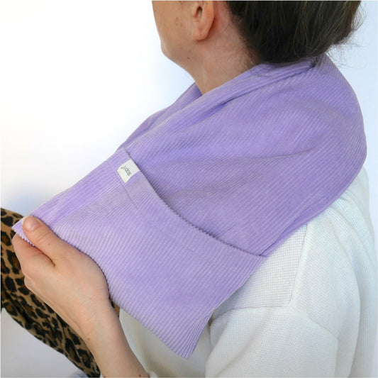 Wärmekissen Hot Cordie Lilac Tree liegt um den Nackenbereich einer Frau, die schräg von hinten im Bild ist. Sie ist mit dem halben Kopf von hinten und bis zur unteren Schulterpartie zu sehen und trägt ein helles Shirt und die Haare zusammengebunden.